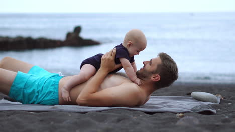 On-the-beach,-a-young-dad-entertains-his-newborn-son-through-playful-interactions.-Embracing-these-precious-moments,-he-finds-joy-in-his-vacation.-His-happiness-radiates-as-a-father